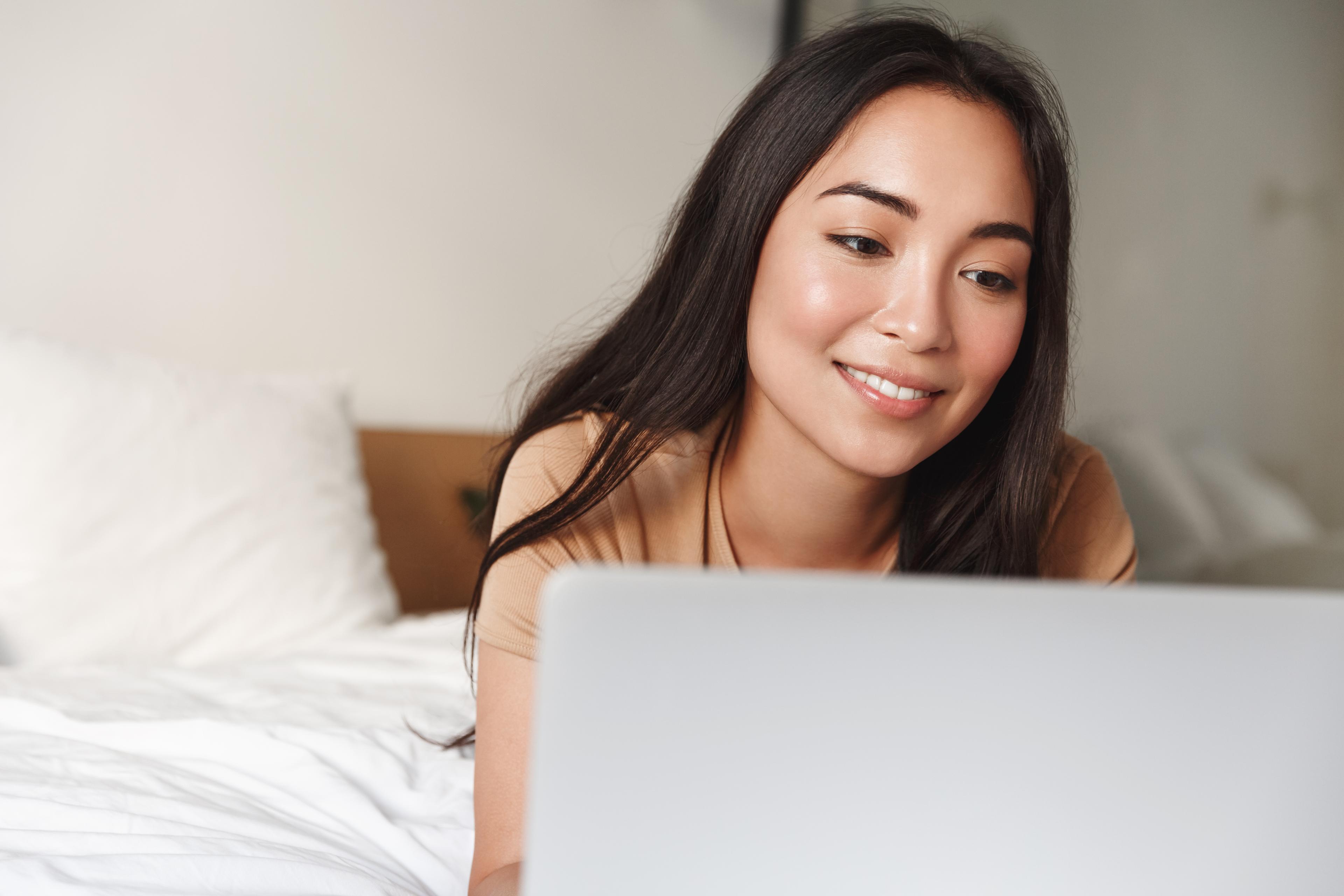 Close-up of girl lying in bed with laptop, smiling at computer screen